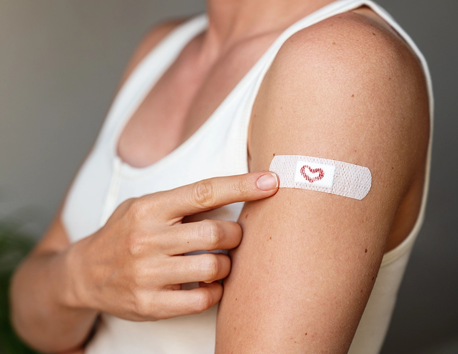 Close-up of a woman in a white tank top pointing to a bandage with a small red heart on her upper arm after receiving a vaccine.